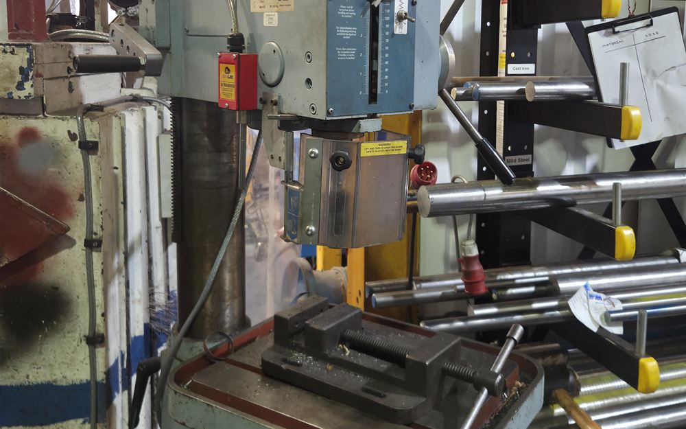 A pillar drill with a machine vice on its table, beside a rack of bar stock.