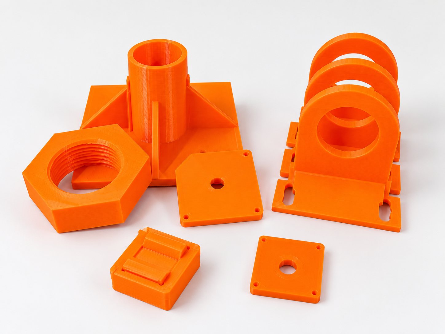A group of orange printed parts on a white surface: a threaded hex nut, a flanged tube on a ribbed base, ring brackets with slotted feet, flat mounting plates and a small open box.